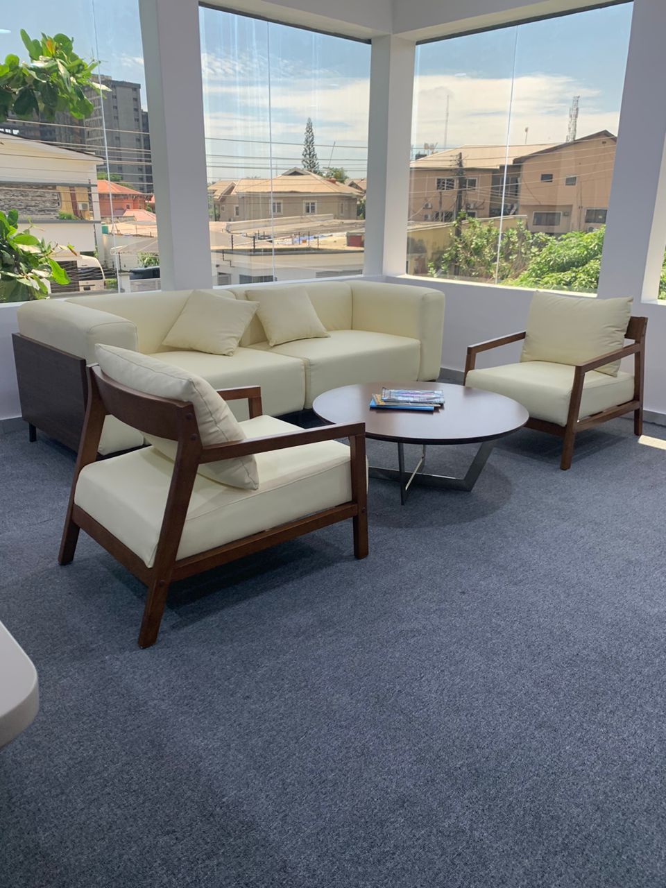 Office seating area with a modern cream-colored sofa, two matching armchairs, and a round wooden coffee table, set against a backdrop of large floor-to-ceiling windows showcasing an urban view